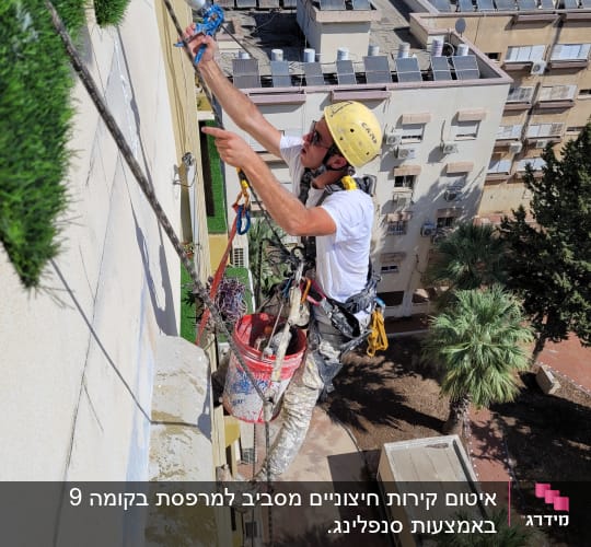 אדם צובע קיר חיצוני בעזרת רתמה ודלי צבע